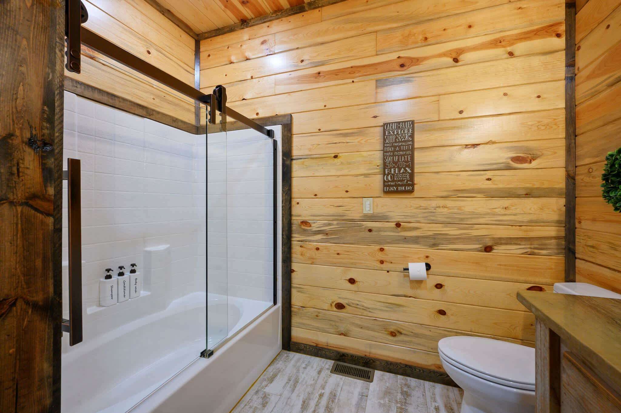 Une salle de bain aux murs lambrissés, dotée d’une douche en verre, de toilettes blanches et d’un meuble-lavabo en bois.
