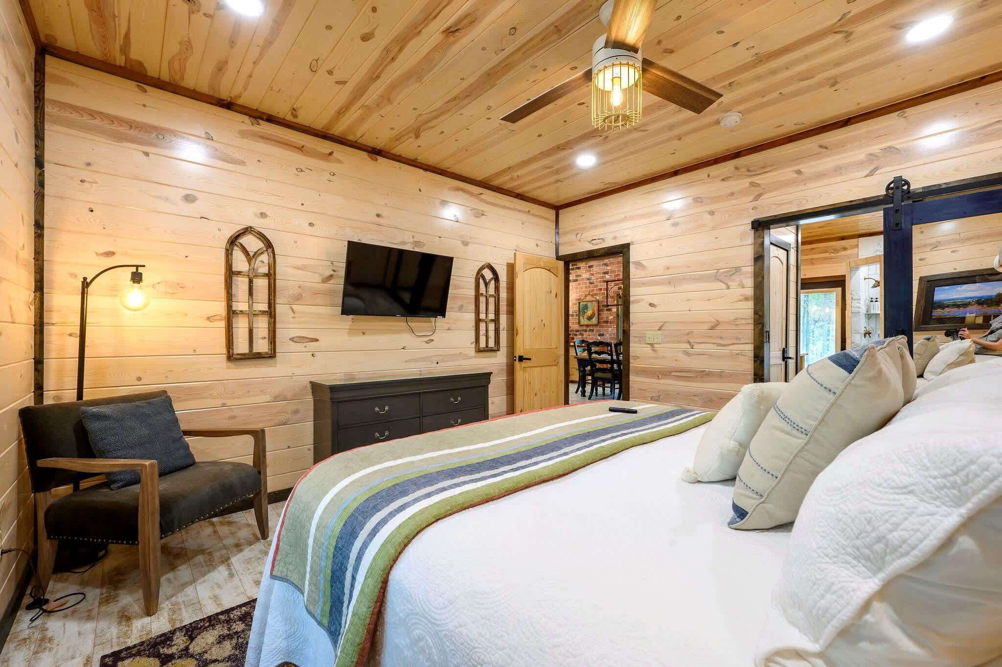 Un dormitorio con paneles de madera, con una cama, una silla, un aparador, un televisor y un ventilador de techo.