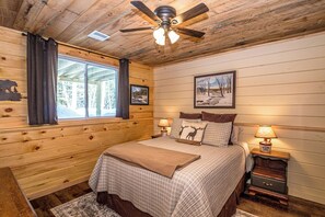 A wooden cabin bedroom with a bed, a nightstand, a lamp, a ceiling fan, and a window with curtains. - Journey's End - 4BR Chalet • 7 Min to Attitash (Bartlett)