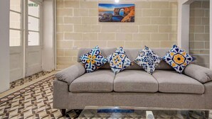 A lobby with a grey sofa, patterned cushions, a tiled floor, a wall-mounted clock, and a framed picture.