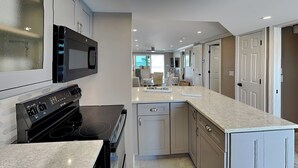 A modern kitchen with a built-in microwave, stove, and dishwasher, a large island with a sink, and a ceiling skylight. - Pointe South 105 | Gulf-Front + Newly Updated (Fort Myers Beach)