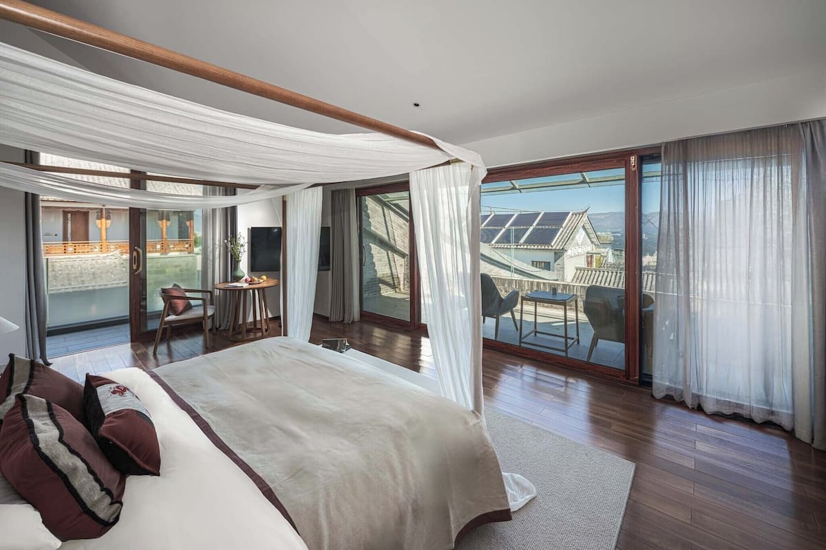 A bedroom with a large bed, a balcony with a table and chairs, and a view of buildings.
