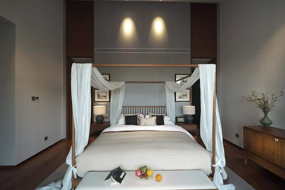 A four-poster bed with sheer curtains, a bedside table with a lamp, and a fruit bowl on the bed.