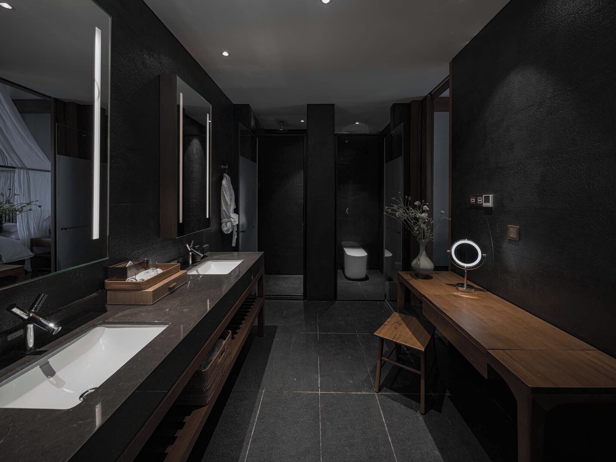 A modern bathroom with dark tiles, a wooden bench, a vanity with a mirror, and a toilet.