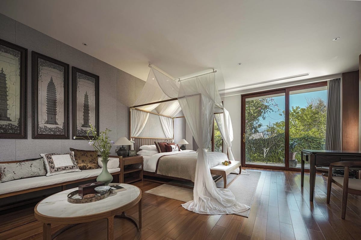 A bedroom with a four-poster bed, a sitting area with a sofa, a round coffee table, and a view through a large sliding glass door.