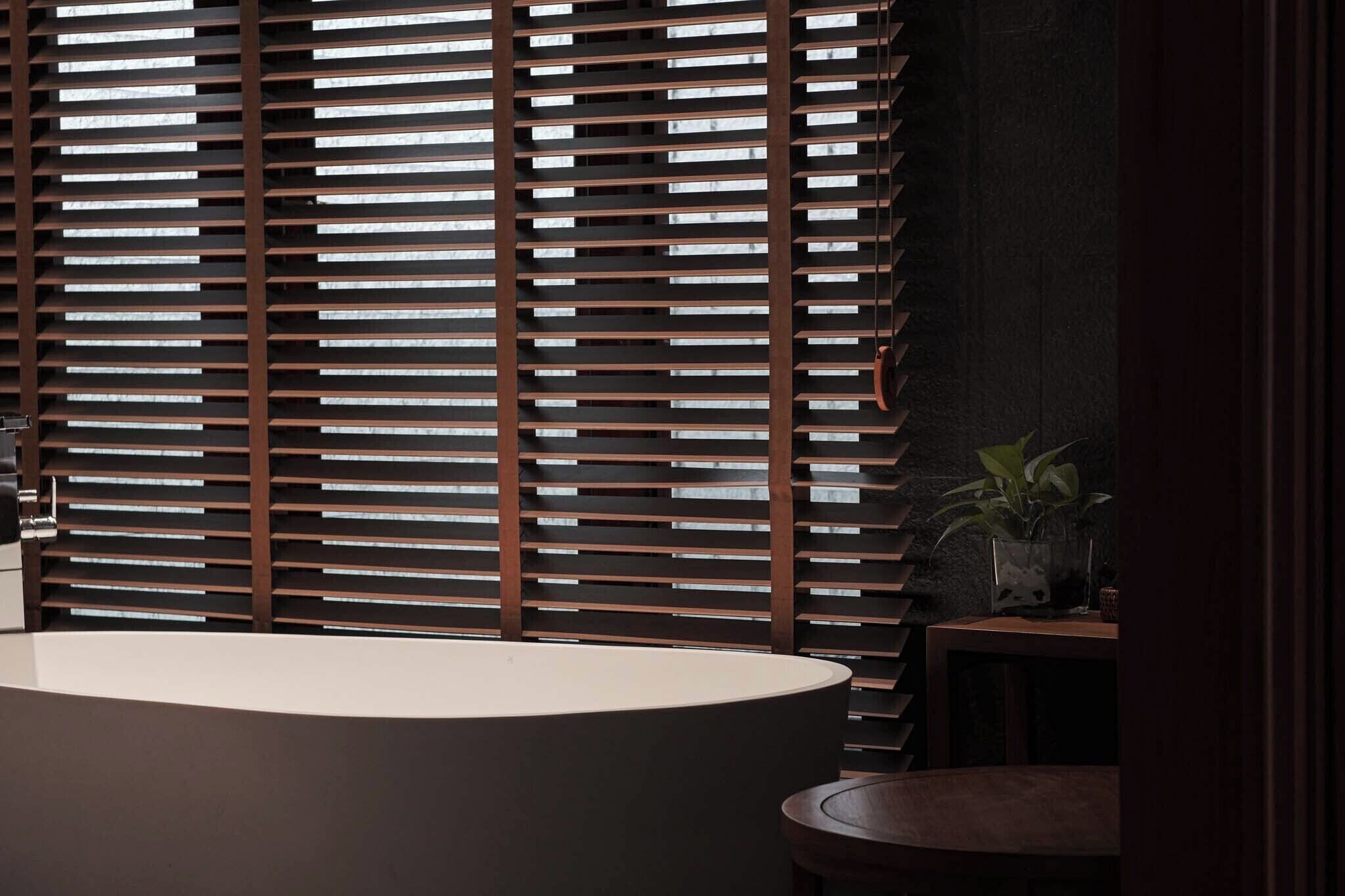 A modern bathroom with a large bathtub, wooden blinds, and a small table with a plant.