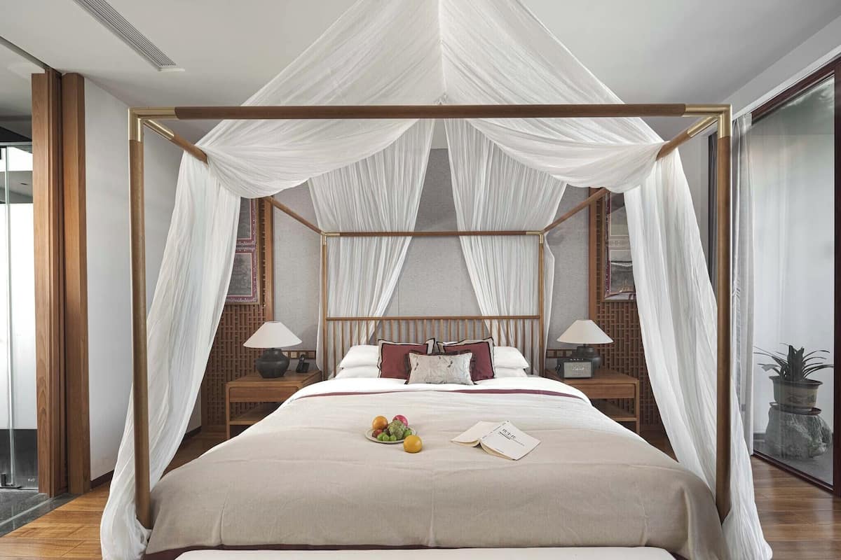 A four-poster canopy bed with a neutral-toned bedding set, bedside lamps, and a fruit plate on the bed.