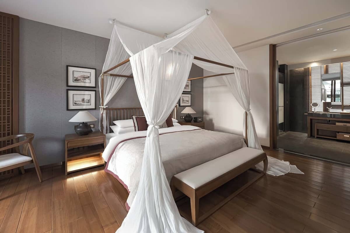 A modern bedroom with a four-poster canopy bed, wooden flooring, bedside tables with lamps, and framed pictures on the wall.