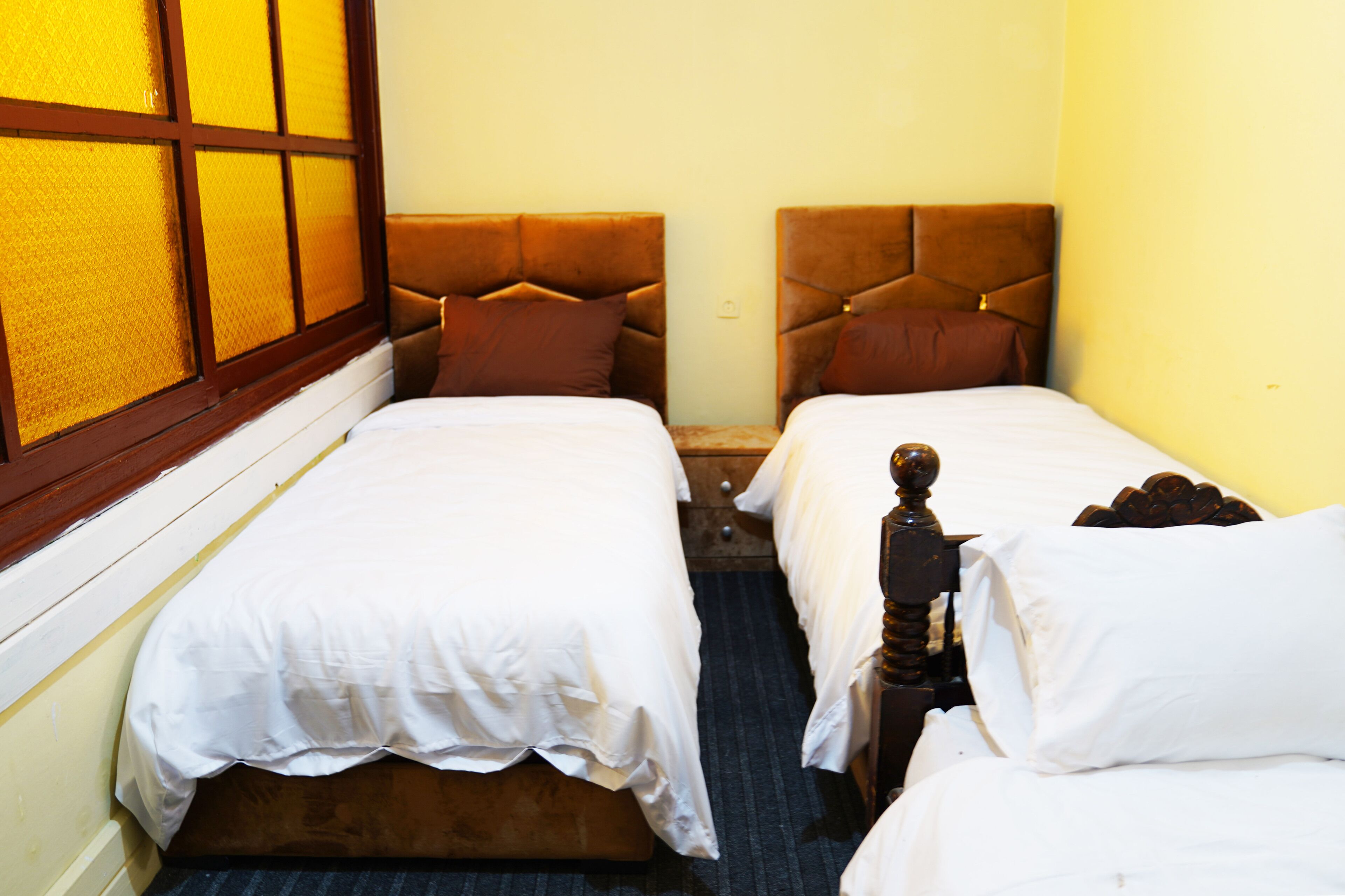 Ein Hotelzimmer mit zwei Einzelbetten, Holz-Kopfteilen, einem Fenster mit gelb getöntem Glas und einem Teppichboden.