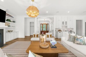 Ein geräumiges Wohnzimmer mit Kamin, einem großen Holztisch, zwei weißen Sofas, einem Essbereich und einer Küche im Hintergrund sowie einer großen Chandelier.