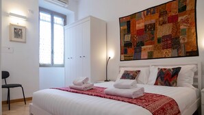 A neatly made bed with white linens and a red patterned throw, a large quilted artwork on the wall, and a white wardrobe. - Casa Sai (Rome)