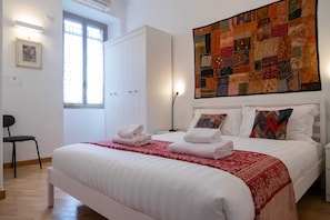 A neatly made bed with white linens and a red patterned throw, a large quilted artwork on the wall, and a white wardrobe. - Casa Sai (Rome)