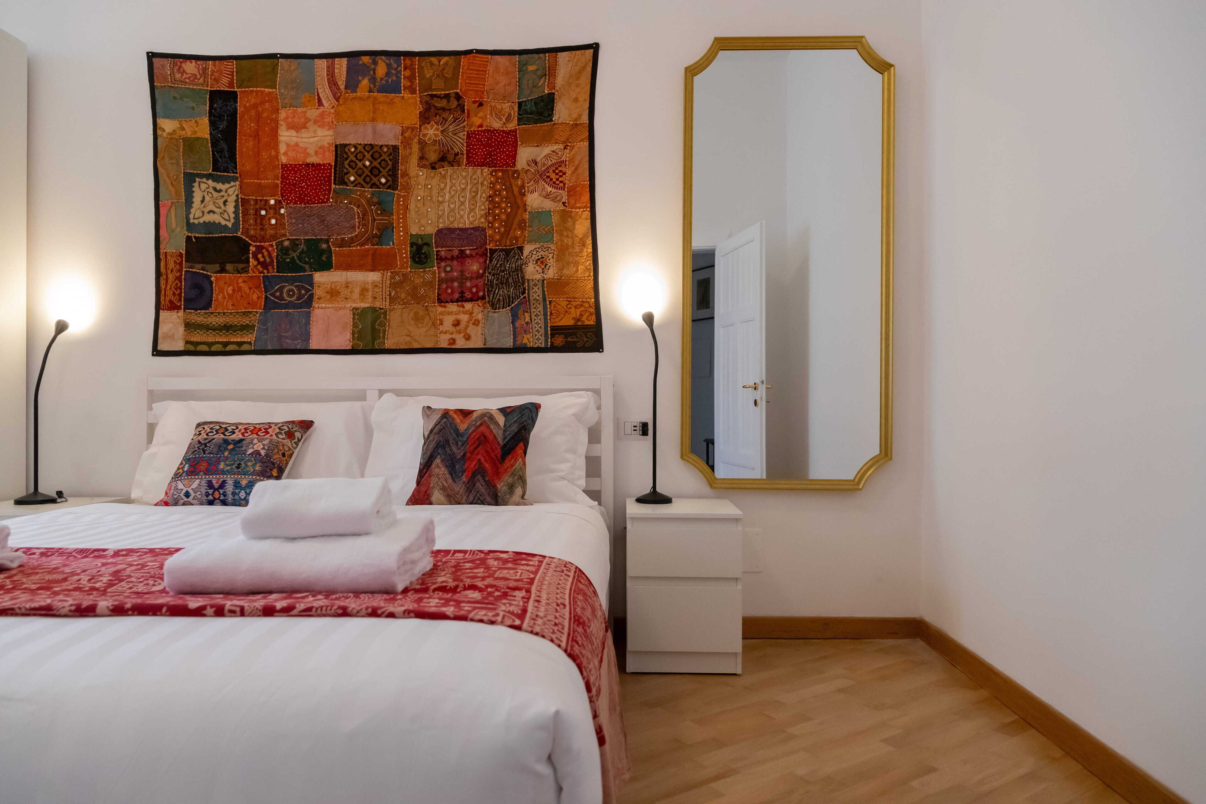 Ein ordentlich gemachtes Bett mit weißen Leinentüchern und einer roten, gemusterten Decke, eine Patchworkdecke mit verschiedenen Stoffresten darüber, ein weißer Nachttisch mit einer Lampe und ein großer Spiegel mit goldener Rahmen an der Wand.