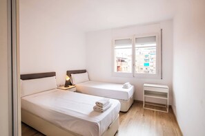 A small, clean room with two beds, a nightstand with a lamp, a window with a view of buildings, and a white shelf. - TUA44PAA1129 - Carrer de Pau Alsina 57 (Barcelona)