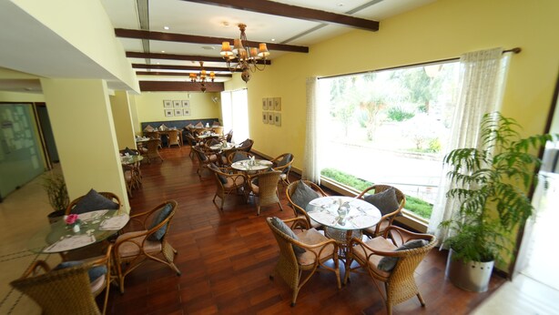 A dining area with wicker furniture and a chandelier, large windows with curtains, and a potted plant.