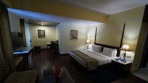 A hotel room with a bed, bedside lamps, a desk, a chair, a TV, and a small table with a vase of flowers.