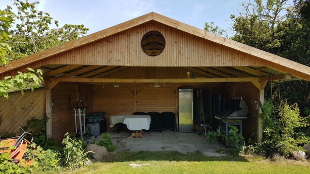 Eine Holzhütte mit einer überdachten Veranda, einem Tisch mit Stühlen, einem Kühlschrank und einem kleinen Tisch mit Gegenständen darauf.