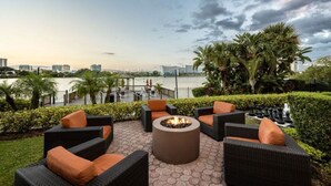 Un área de estar al aire libre con muebles de mimbre y un hogar central, con vistas a un cuerpo de agua y al horizonte urbano.