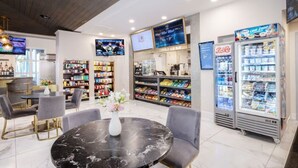 Un café moderno con una mesa de mármol, sillas grises, un refrigerador, estantes llenos de aperitivos y una pantalla de televisión.