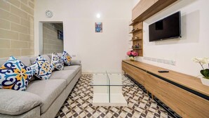 A living room with a sofa, patterned cushions, a glass coffee table, a wooden console table, a TV, and a wall clock. - Private Room in Hamrun with Shared Kitchen (Hamrun)