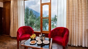 Ein Zimmer mit zwei roten Samtstühlen, einem Holztisch mit Frühstücksgebäck und einem großen Fenster mit Blick auf Grünflächen und Berge.