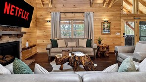 A log cabin interior with a fireplace, a sofa, a coffee table, and a television displaying 'NETFLIX'. - Family-friendly cabin with game room and backyard! (Hanover)
