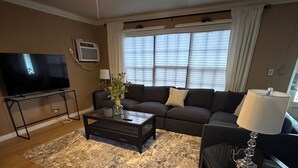 Una sala de estar con ventilador de techo, televisor sobre un mueble bajo, sofá, mesa de centro, lámpara y ventana con persianas.
