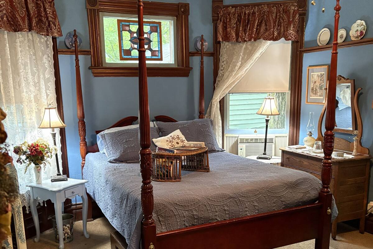 A four-poster bed in a room with blue walls, a window with a view, and a small bedside table with a lamp.