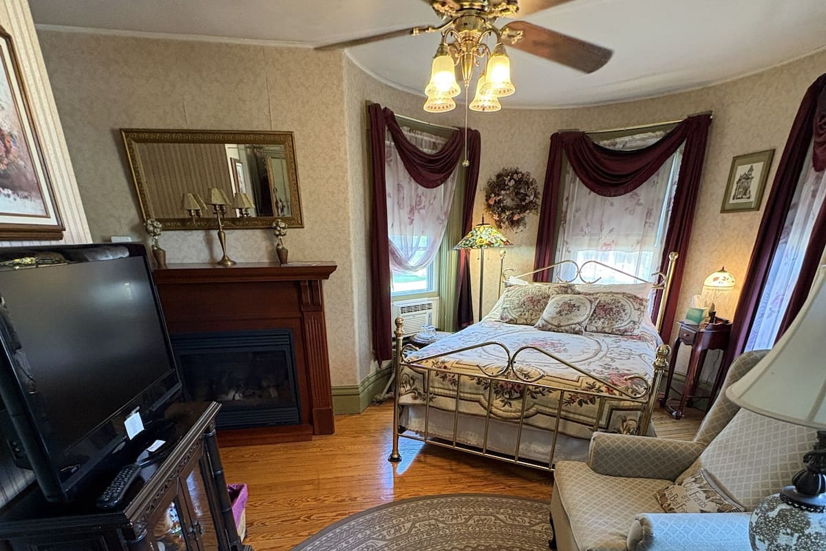 A room with a bed, a sofa, a television, a fireplace, and a ceiling fan.