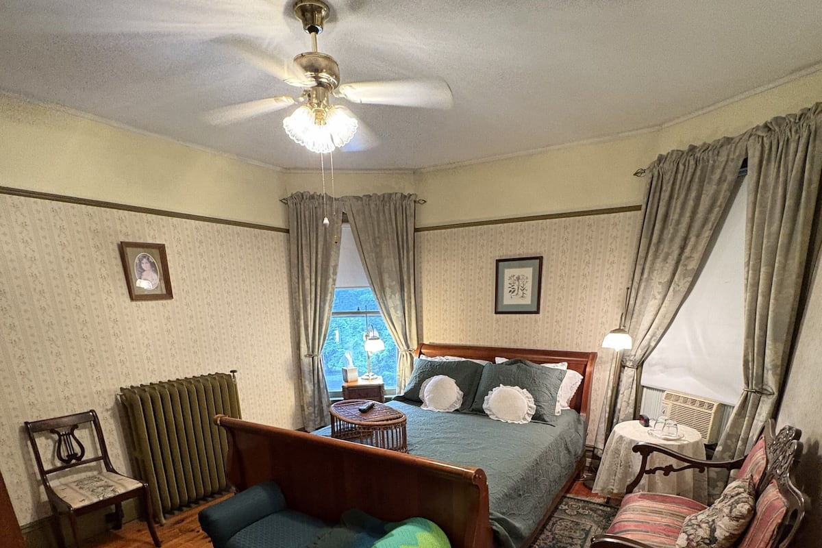 A bedroom with a bed, a chair, a sofa, a ceiling fan, and a window.