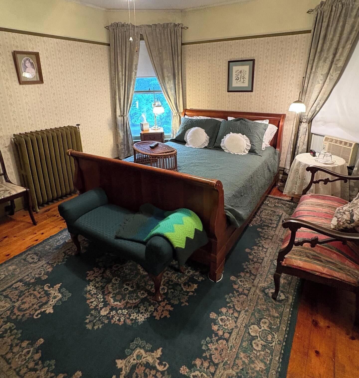 A bedroom with a bed, a sofa, a table, a chair, a radiator, and a window with curtains.