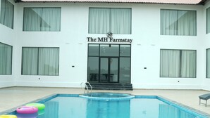 A swimming pool with a few inflatable rings, a building with a sign reading 'The MH Farmstay,' and large windows. - THE MH Farmstay Hyderabad | Luxury Pool Villa for Parties & Getaways (Hyderabad)