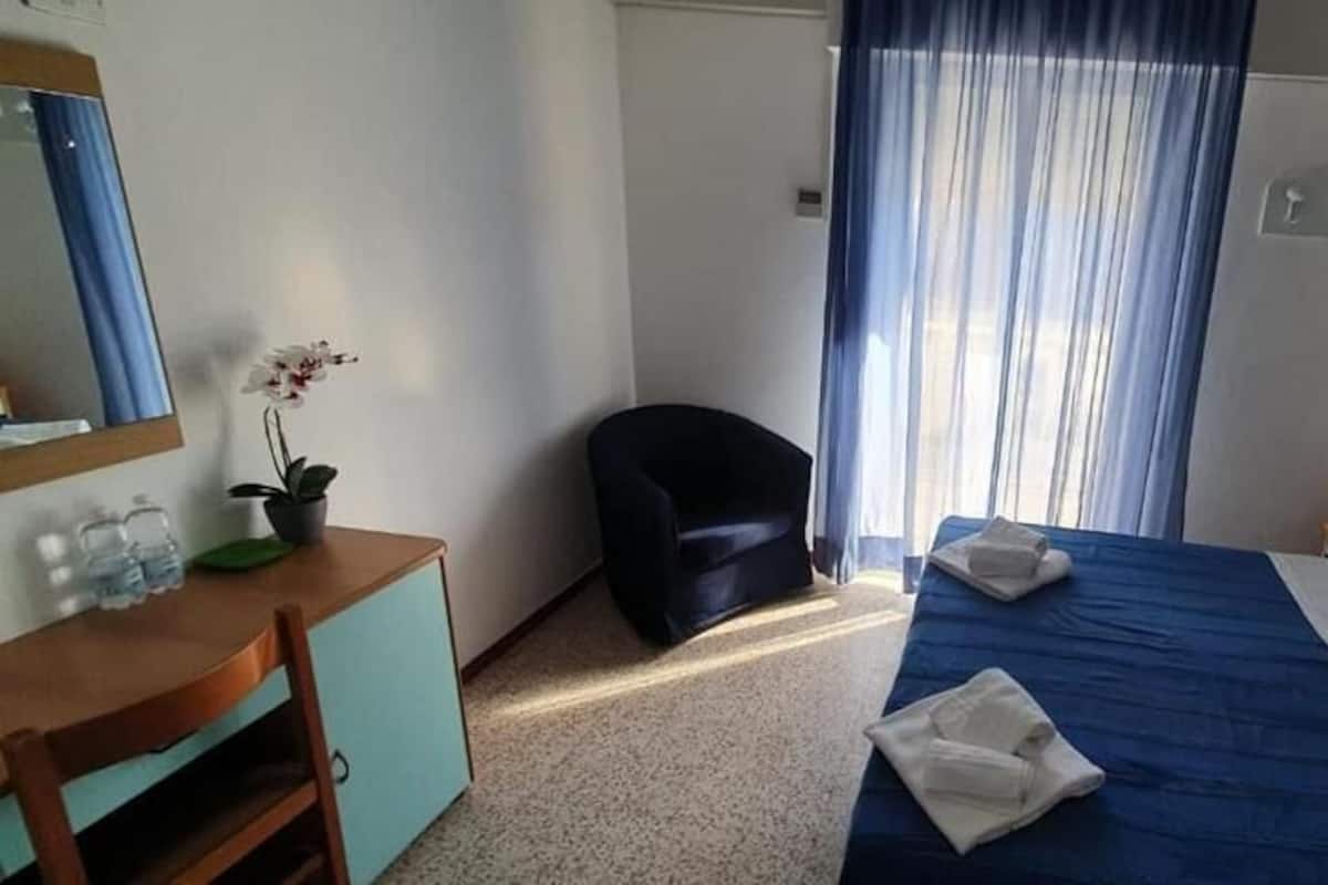 Ein Hotelzimmer mit einem Bett, einem Schreibtisch mit Stuhl, einem kleinen Tisch mit Blumentopf und einem Fenster mit Vorhängen.