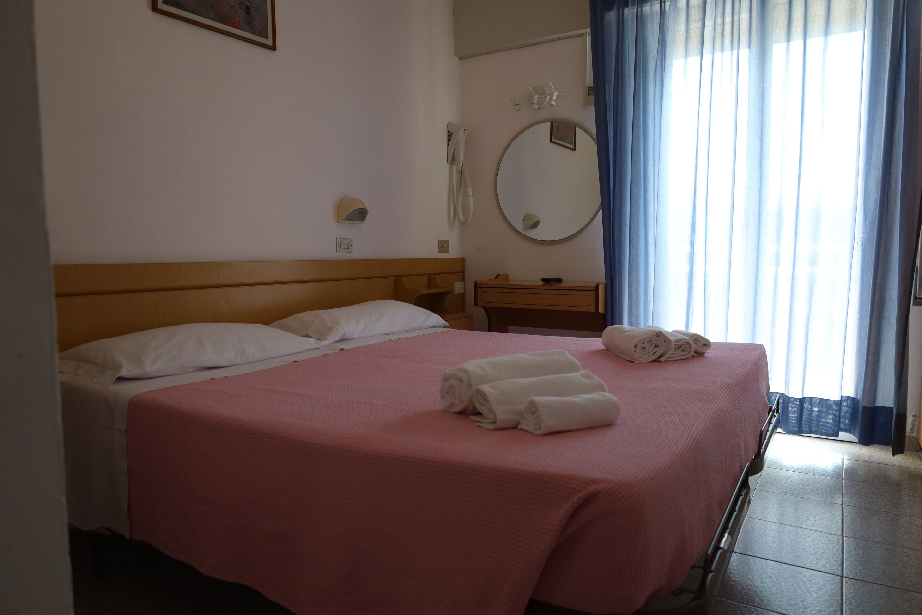 A hotel room with a bed, a bedside table, a mirror, and a window with curtains.