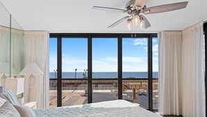 A bedroom with a large bed, a ceiling fan, and a view of the beach through large windows. - Golden Sands 104 by Seaside Vacations & Sales (Ocean City)
