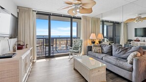 A spacious living room with a balcony, a large ceiling fan, and a television. - Golden Sands 104 by Seaside Vacations & Sales (Ocean City)