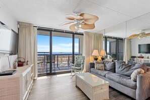 A spacious living room with a balcony, a large ceiling fan, and a television. - Golden Sands 104 by Seaside Vacations & Sales (Ocean City)