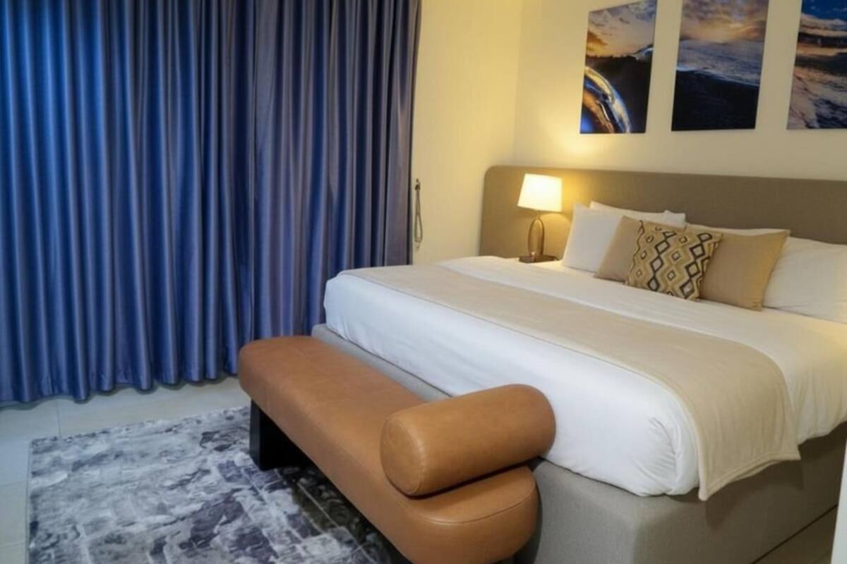 A hotel room with a bed, a bench, a lamp, and curtains.