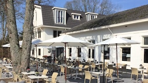 An outdoor seating area with tables and chairs, a building with a sloped roof, and trees in the background. - Explore the Dutch Outdoors Without Compromising Comfort! Free Parking, Dining! (Beekbergen)