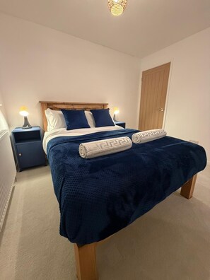A neatly made bed with a blue blanket and two pillows, a wooden headboard, and a nightstand with a lamp. - Luxury Home|Coffee Machine| En-suite| (Redditch)