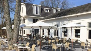 An outdoor seating area with tables and chairs, a building with a sloped roof, and trees in the background. - From Adventure Parks to Art Museums—Stay Near It All! Terrace, Onsite Dining! (Beekbergen)