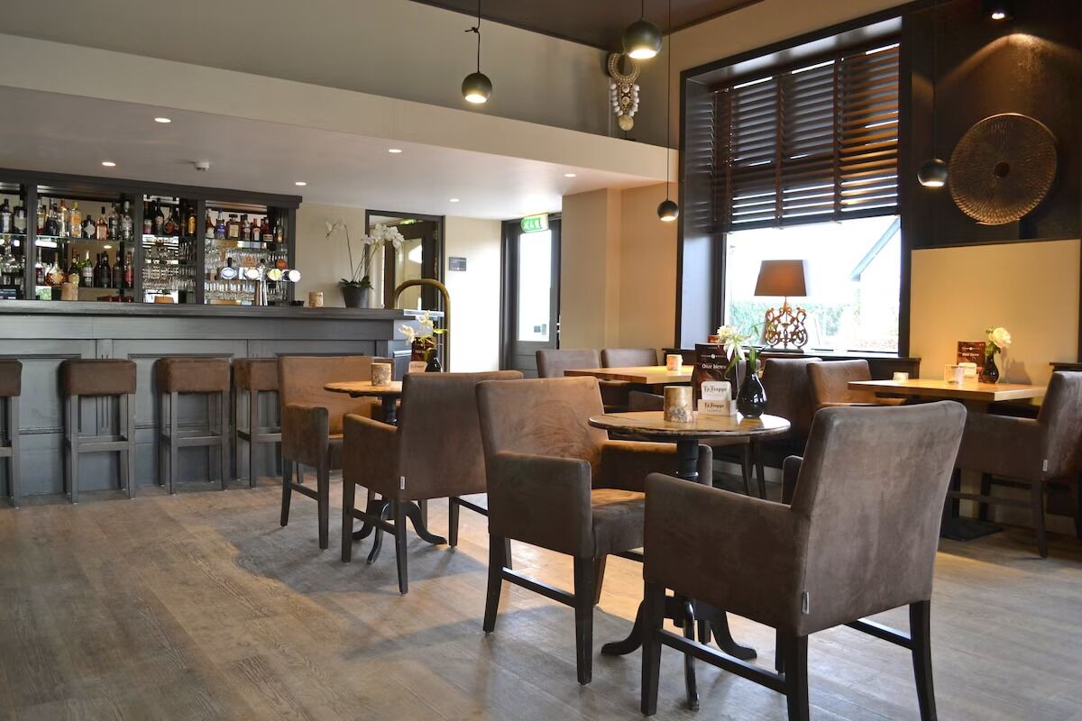 A restaurant with a bar area, wooden tables and chairs, and a large window with blinds.