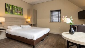 Hotelkamer met een bed, bureau, stoel, televisie, raam met zonwering, schilderij van een hert en een vaas met bloemen.