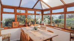A conservatory with a wooden table and chairs, large windows, and a view of greenery. - 2 Oak Tree Cottages (Axminster)