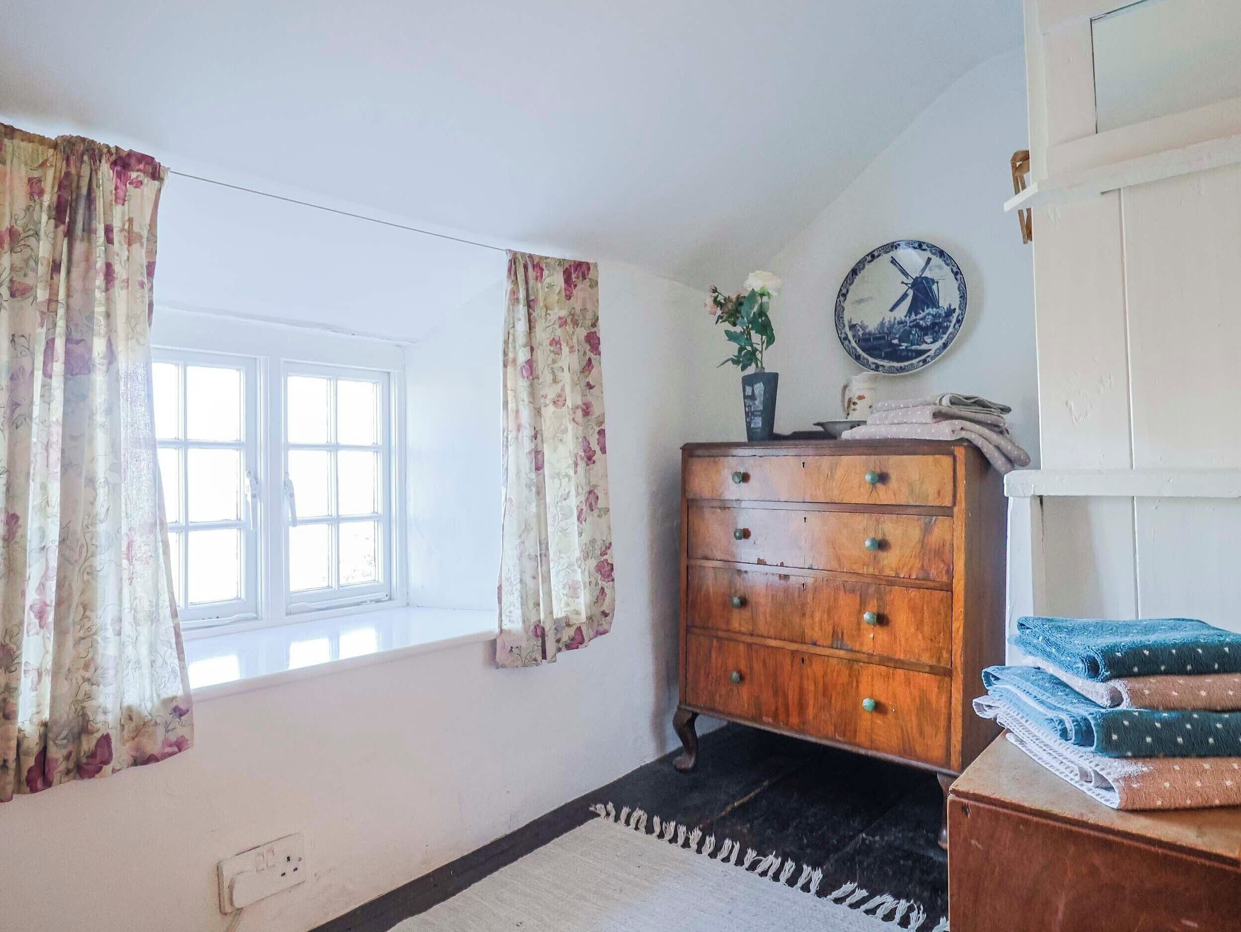 A room with a window, a wooden dresser, a clock, and a wall-mounted plate.