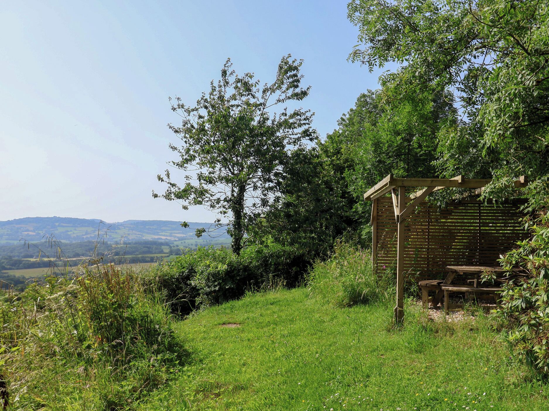 A wooden gazebo with a bench, surrounded by lush greenery and a view of distant hills.