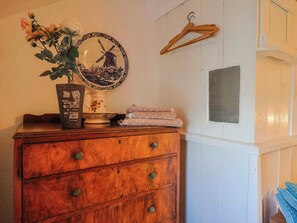 Eine Holzkommode mit Schubladen, eine Vase mit Blumen, ein Teller mit Windmühlenmotiv und ein Holzaufhänger an der Wand.