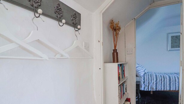 A narrow hallway with a white coat rack, a white bookshelf with books, and a bed with a striped blanket. - 2 Oak Tree Cottages (Axminster)