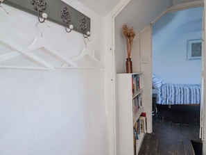 A narrow hallway with a white coat rack, a white bookshelf with books, and a bed with a striped blanket. - 2 Oak Tree Cottages (Axminster)
