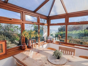 A conservatory with a wooden table and chairs, surrounded by large windows offering a view of the garden. - 2 Oak Tree Cottages (Axminster)